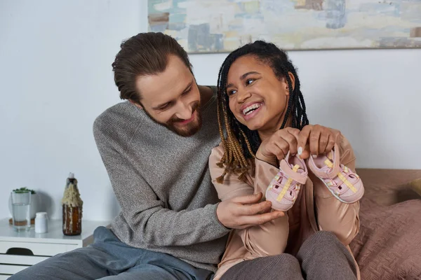 Feliz mulher americana africana segurando minúsculos sapatos de bebê perto do marido, futuros pais, expectativa, fofa — Fotografia de Stock