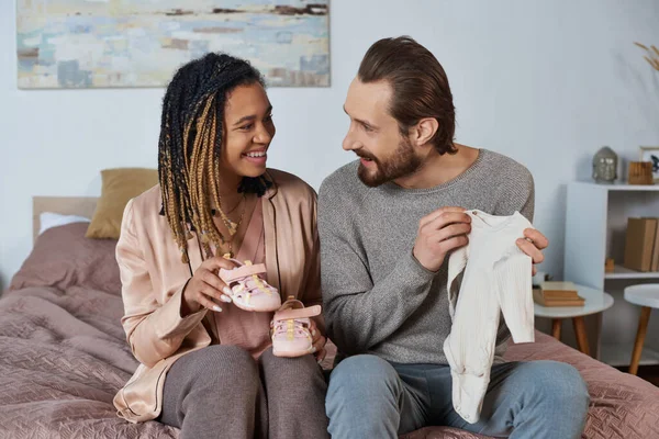 Feliz casal interracial segurando roupas de bebê bonito, sapatos minúsculos, futuros pais, expectativa — Fotografia de Stock
