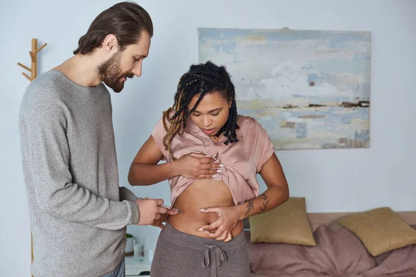 Bearded man making hormonal injections in belly of african american wife, tattoo, fertility, couple — Stock Photo