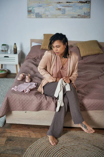 Femme afro-américaine tatouée tenant des vêtements de bébé, assise sur le lit, bouleversée, concept de fausse couche — Photo de stock