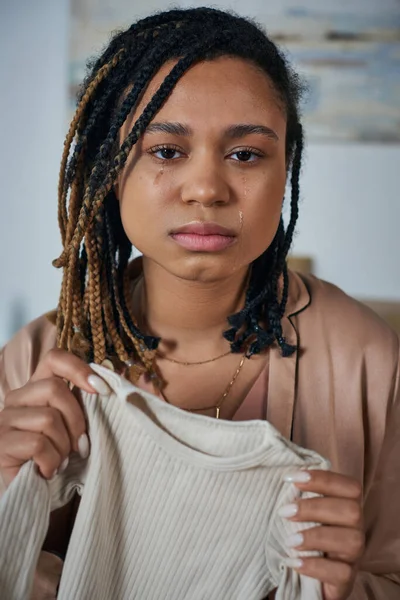 Donna africana americana depressa che tiene i vestiti del bambino e piange a casa, concetto di aborto spontaneo — Foto stock