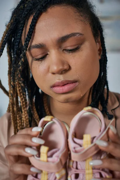 Sad african american woman holding baby clothes and crying at home, miscarriage concept, close up — Stock Photo