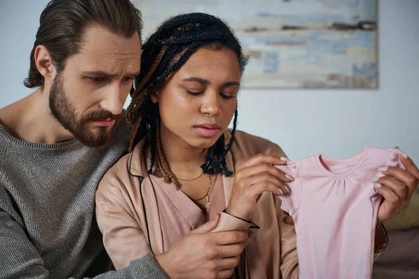 Triste hombre calmando afroamericana esposa llorando y sosteniendo bebé ropa, aborto espontáneo concepto - foto de stock