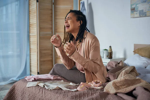 Deuil, femme afro-américaine déprimée pleurant près des vêtements de bébé sur le lit, concept de fausse couche — Photo de stock