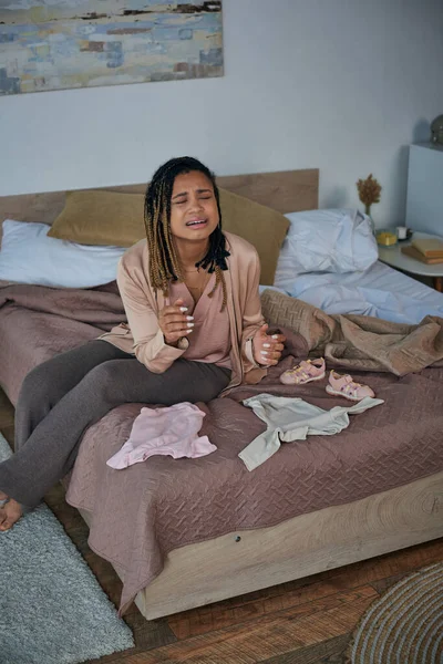 Desesperación, mujer afroamericana deprimida llorando cerca de ropa de bebé en la cama, concepto de aborto espontáneo - foto de stock