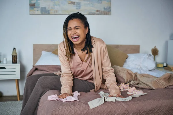 Disperazione, donna afro-americana che piange vicino a vestiti per bambini sul letto, concetto di aborto spontaneo, depressione — Foto stock