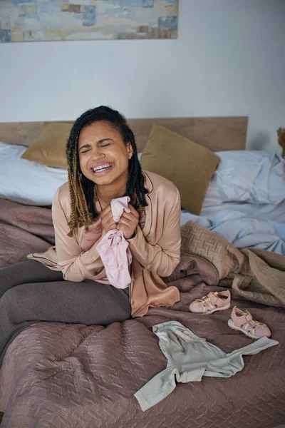 Dolore, donna afro-americana che piange vicino a vestiti per bambini sul letto, concetto di aborto spontaneo, depressione — Foto stock