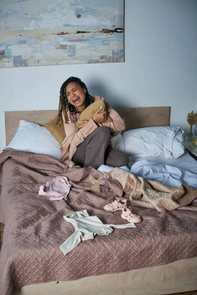 Dolore, donna afro-americana che tiene il cuscino e piange vicino ai vestiti per bambini sul letto, aborto spontaneo — Foto stock