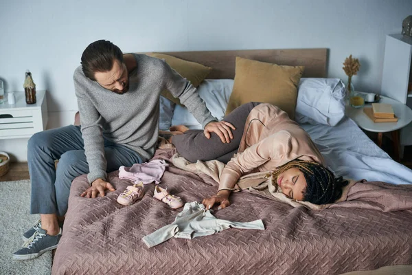 Deuil, concept de fausse couche, homme apaisant femme afro-américaine pleurant près des vêtements de bébé, couché sur le lit — Photo de stock