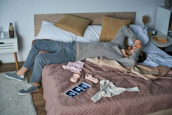 Trauer, depressiver Mann liegt im Bett neben Babykleidung und Ultraschalluntersuchung, Fehlgeburtenkonzept — Stockfoto