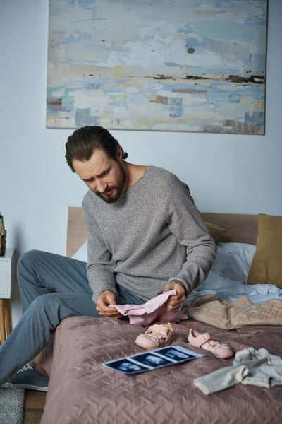 Loss, depressed man sitting on bed near baby clothes and ultrasound scan, miscarriage concept — Stock Photo