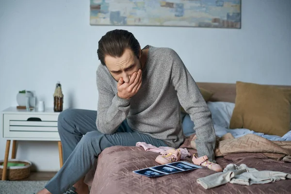 Ungläubiger, depressiver Mann sitzt auf Bett neben Babykleidung und Ultraschalluntersuchung, Fehlgeburtenkonzept — Stockfoto