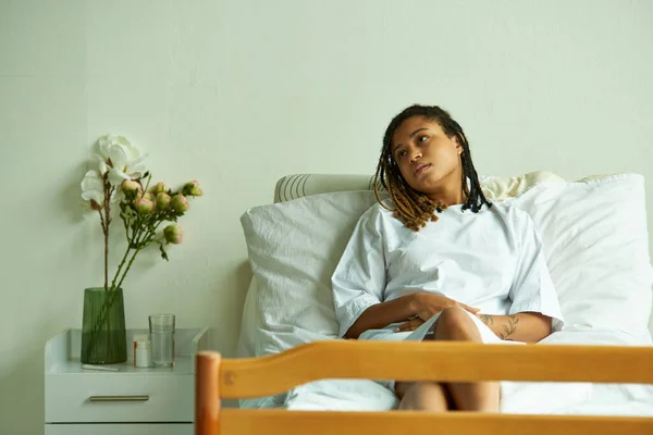 Triste mulher afro-americana deitado na ala privada, flores, vidro de água, hospital, aborto espontâneo — Fotografia de Stock