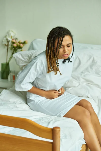 Mujer afroamericana sentada en la cama, tocando el vientre, sala privada, hospital, concepto de aborto espontáneo - foto de stock