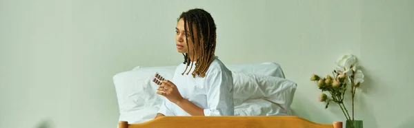 Mujer afroamericana en bata de hospital sosteniendo pastillas, sala privada, paciente en clínica, pancarta - foto de stock