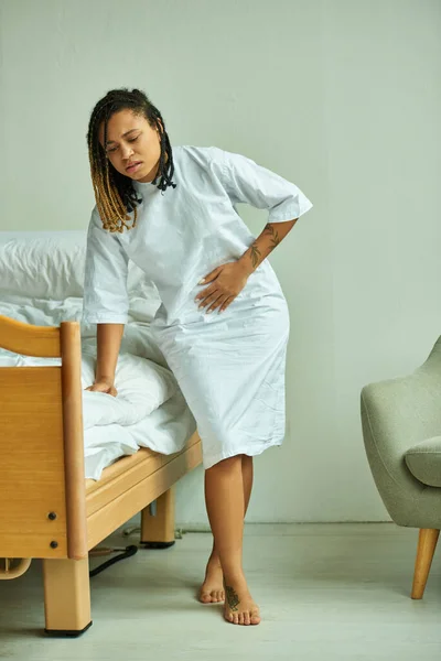 Donna afroamericana tatuata in abito ospedaliero che tocca pancia, reparto privato, paziente in clinica — Foto stock