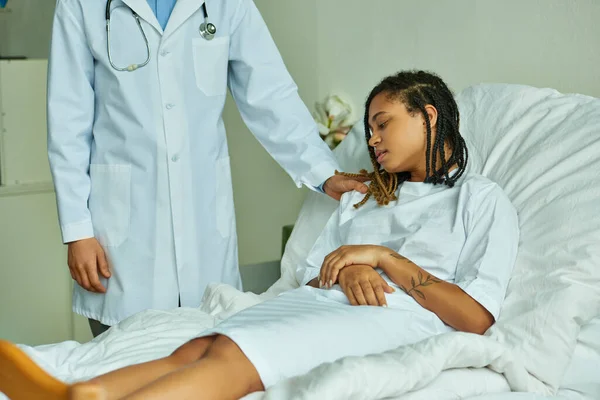 Doctor barbudo calmando mujer afroamericana en bata de hospital, sala privada, paciente - foto de stock