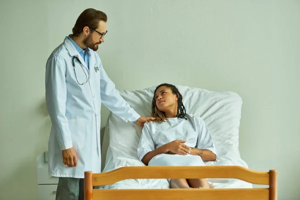 Médecin dans des lunettes debout près de femme afro-américaine en robe d'hôpital, salle privée, patient — Photo de stock