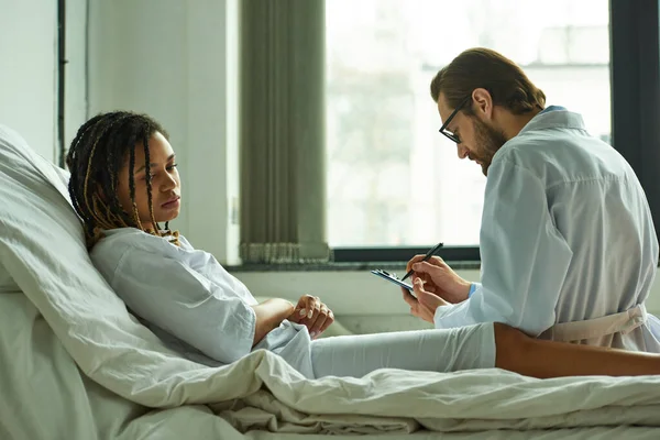 Doctor in glasses writing prescription to upset african american woman, private ward, hospital — Stock Photo