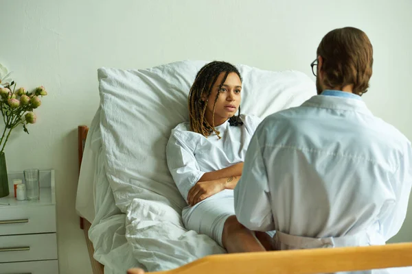 Doutor no revestimento branco que aconselha a mulher americana africana, enfermaria privada, hospital, assistência médica — Fotografia de Stock