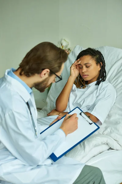 Doctor in glasses writing prescription to worried african american woman, private ward, hospital — Stock Photo