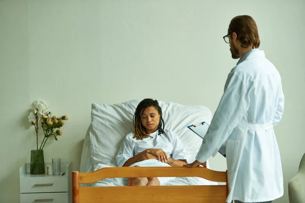Doctor de pie con portapapeles cerca de la mujer afroamericana, sala privada, hospital, consulta - foto de stock
