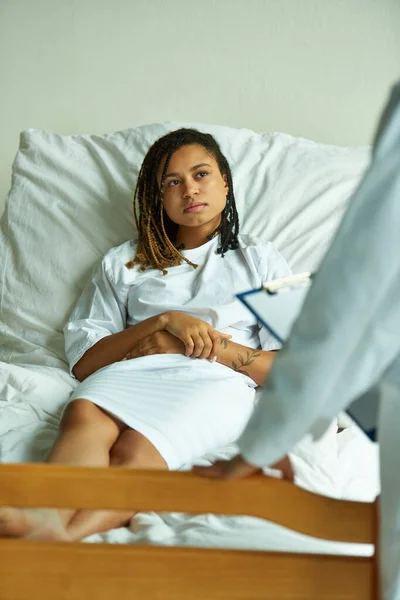 Médico de pie con portapapeles cerca de la mujer afroamericana, sala privada, consulta en la clínica - foto de stock