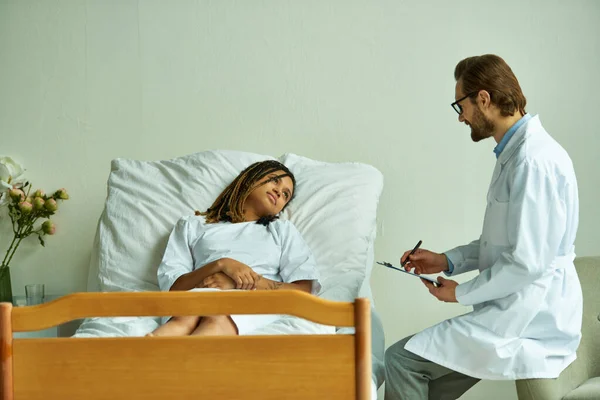 Bearded doctor standing with clipboard near african american woman, private ward, consult — Stock Photo