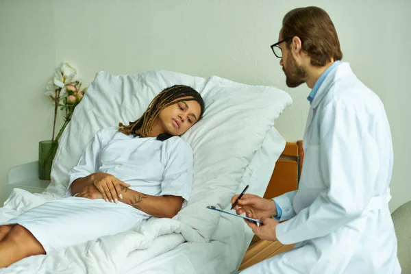 Bearded doctor with clipboard standing near sad african american woman, private ward, consult — Stock Photo