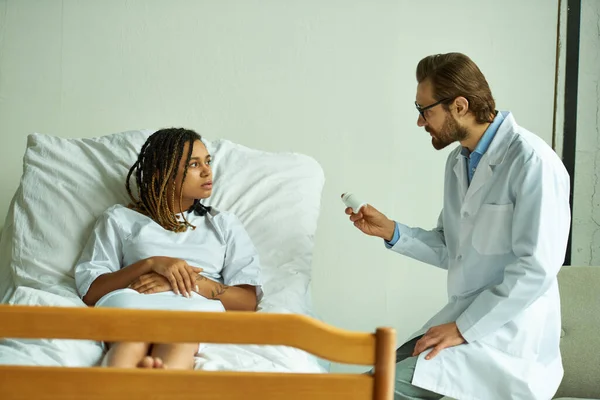 Médico segurando garrafa com pílulas perto de mulher americana africana, enfermaria privada, prescrevendo medicação — Fotografia de Stock