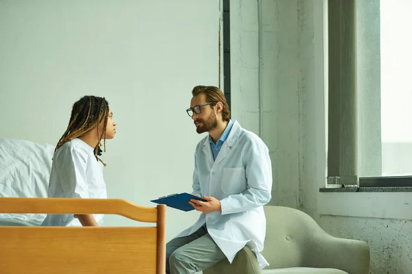 Docteur en lunettes et manteau blanc tenant presse-papiers, parlant à une femme afro-américaine, salle privée — Photo de stock