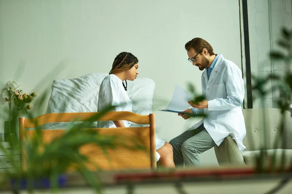 Médecin en lunettes et manteau blanc tenant presse-papiers, patient afro-américain, salle privée — Photo de stock