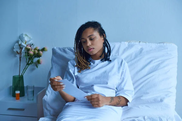 Частная палата, грустная африканская американка в больничном халате, рассматривает ультразвук, концепцию выкидыша — стоковое фото