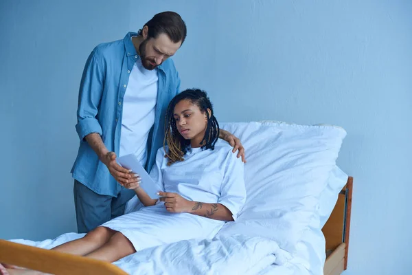 Mann steht neben trauriger afrikanisch-amerikanischer Frau, schaut Ultraschall, Krankenhaus, Fehlgeburtenkonzept — Stockfoto