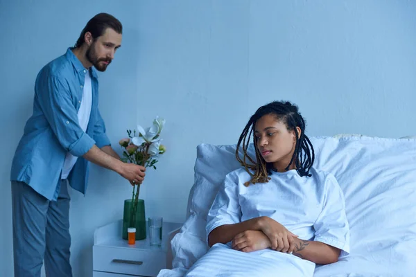 Conceito de aborto espontâneo, homem colocando flores em vaso perto de esposa afro-americana deprimida, enfermaria hospital — Fotografia de Stock