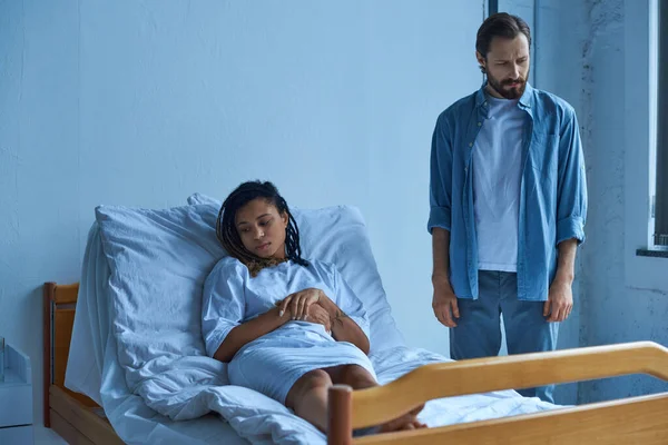 Концепція викидня, сумна афроамериканка сумує, лежачи в лікарняному ліжку біля чоловіка, підопічна — стокове фото