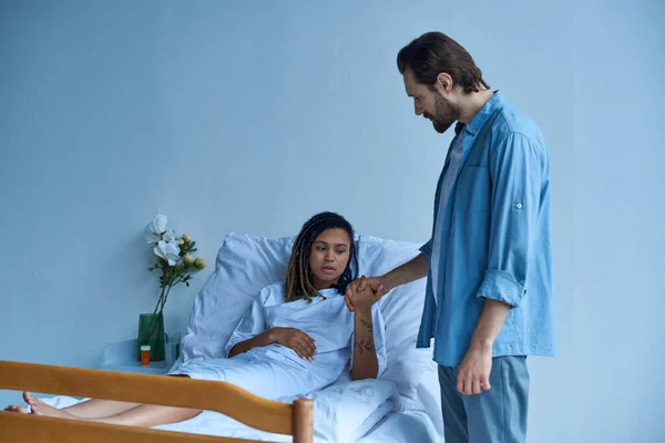 Aborto espontâneo, homem de mãos dadas de triste esposa afro-americana, dor, cama de hospital, enfermaria privada — Fotografia de Stock