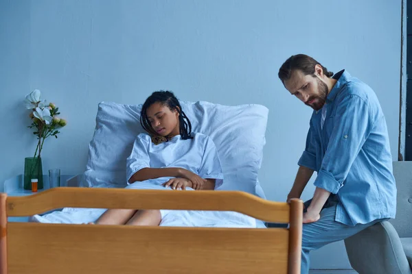 Aborto espontáneo, hombre sentado cerca de la deprimida esposa afroamericana, dolor, cama de hospital, sala privada - foto de stock