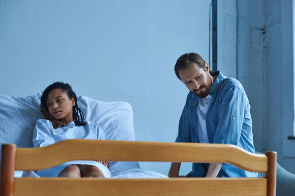 Aborto espontáneo, hombre sentado cerca de la mujer afroamericana deprimida, dolor, cama de hospital, sala privada - foto de stock