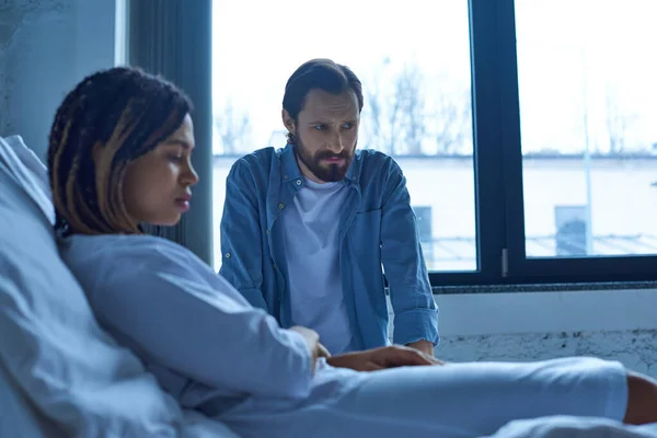 Aborto espontâneo, homem sentado perto de mulher afro-americana deprimida, borrão, cama de hospital, enfermaria privada — Fotografia de Stock