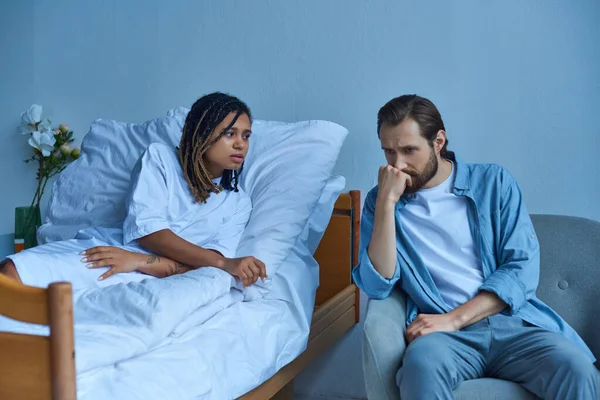Hombre deprimido sentado cerca de esposa afroamericana enferma, hospital, sala privada, dolor, clínica - foto de stock