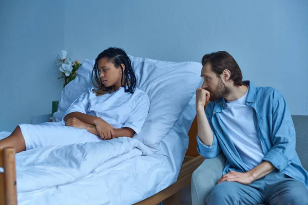 Hombre afligido cerca de la deprimida esposa afroamericana, sala de hospital, concepto de aborto espontáneo, desesperación - foto de stock