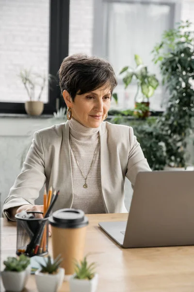 Досвідчена бізнес-леді біля ноутбука і напій на робочому місці в сучасному офісі — стокове фото