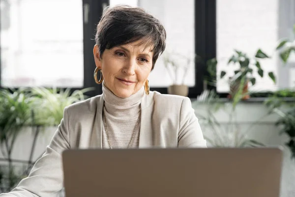 Позитивна бізнес-леді середнього віку, що працює біля ноутбука в сучасному офісі, успіх бізнесу — стокове фото