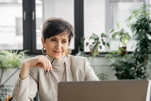 Mulher de negócios madura segurando caneta e pensando perto de laptop no escritório, tomada de decisão, planejamento — Fotografia de Stock