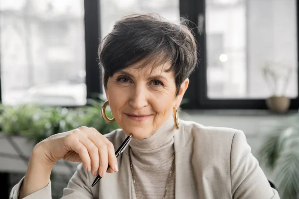 Mature woman entrepreneur with pen looking at camera in office, experience, professional headshot — Stock Photo