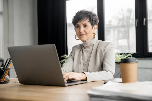 Femme entrepreneur mature tapant sur ordinateur portable près à emporter boisson dans un environnement de travail moderne — Photo de stock