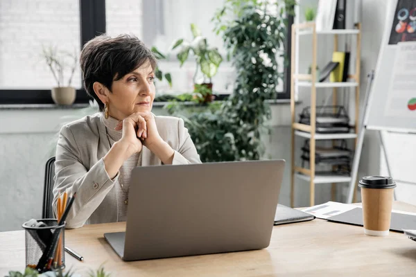 Donna d'affari premurosa seduta vicino al computer portatile e guardando altrove in ufficio moderno, esecutivo, ambizioso — Foto stock