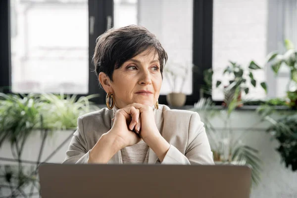 Thoughtful mature businesswoman entrepreneur looking away near laptop in office — Stock Photo