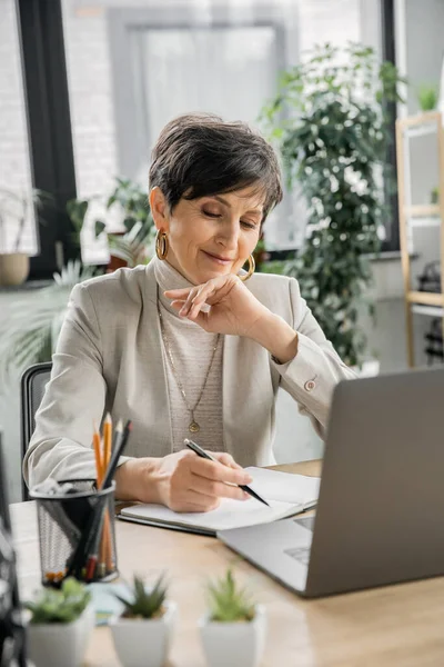 Smiling middle aged businesswoman writing in notebook near laptop in modern office space — Stock Photo